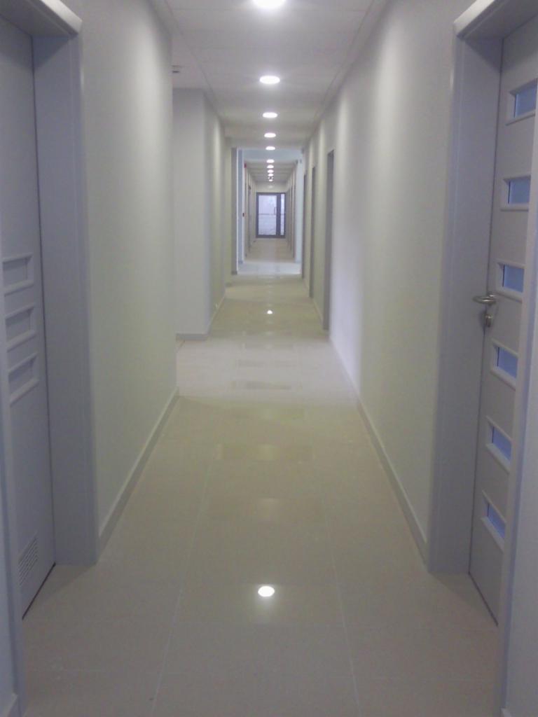 Długi, jasny korytarz z białymi ścianami, wykończony błyszczącymi, kremowymi płytkami podłogowymi i rzędem wbudowanych lamp sufitowych, z widocznymi fragmentami białych drzwi po bokach.