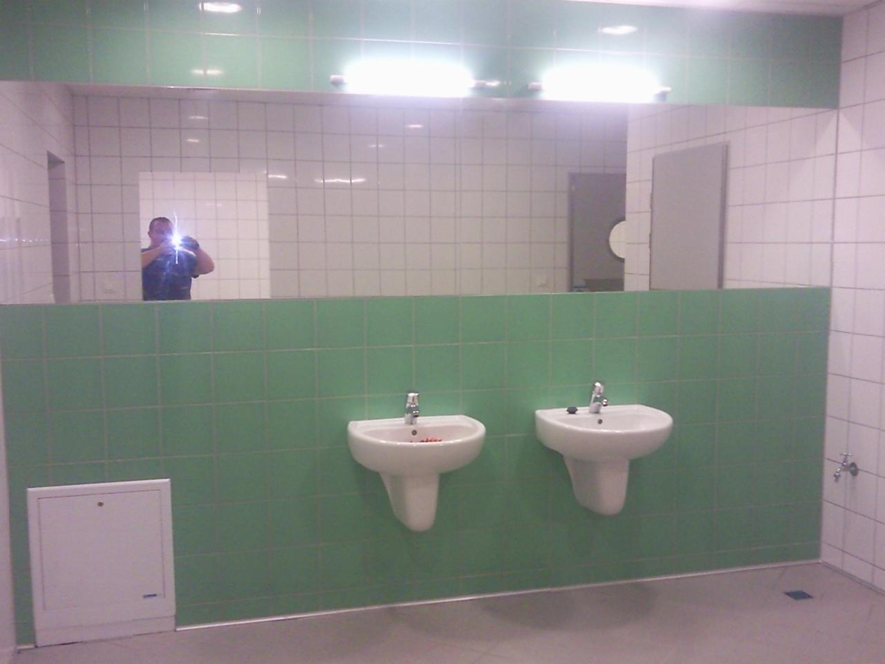 Łazienka publiczna z dwoma umywalkami na tle zielonych i białych płytek, z odbiciem osoby robiącej zdjęcie w lustrze.
