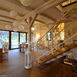 Jasne wnętrze sali z drewnianą podłogą, stołami, krzesłami, dekoracyjnymi lampionami, schodami z ozdobną balustradą i kącikiem zabaw dla dzieci.