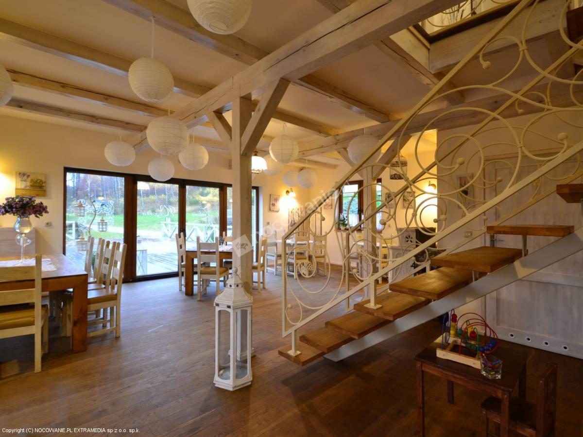 Jasne wnętrze sali z drewnianą podłogą, stołami, krzesłami, dekoracyjnymi lampionami, schodami z ozdobną balustradą i kącikiem zabaw dla dzieci.