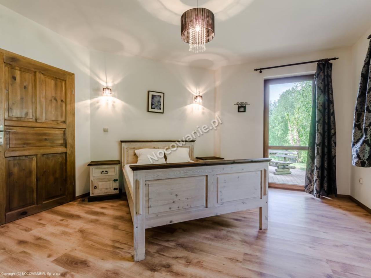 Jasne wnętrze sypialni z łóżkiem o drewnianej ramie, widokiem na taras i ogrodem za oknem, oświetlone lampami ściennymi i żyrandolem.