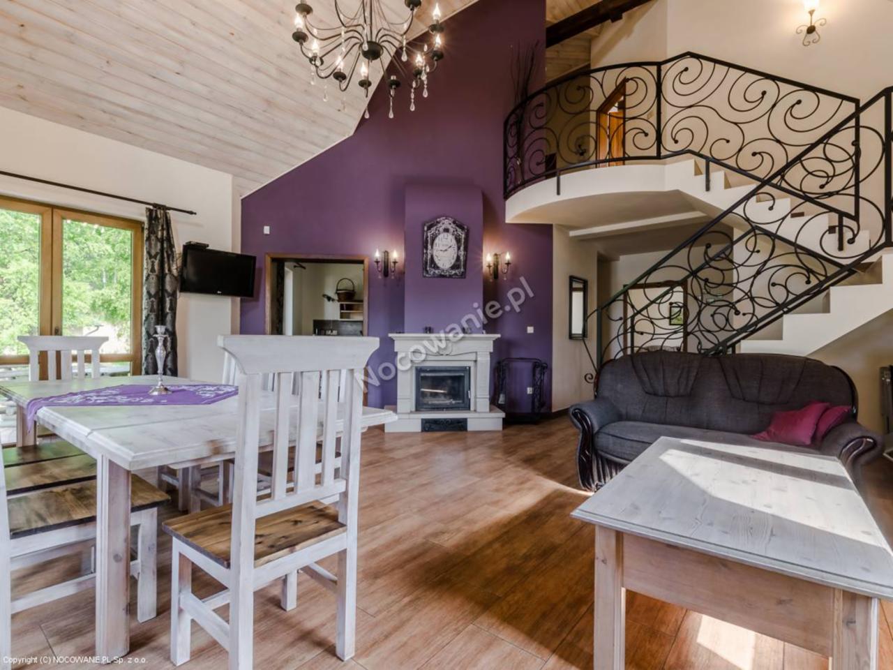 Przestronne wnętrze domu z drewnianą podłogą, białym stołem i krzesłami, kominkiem, fioletową ścianą i schodami z ozdobną, kutą balustradą.