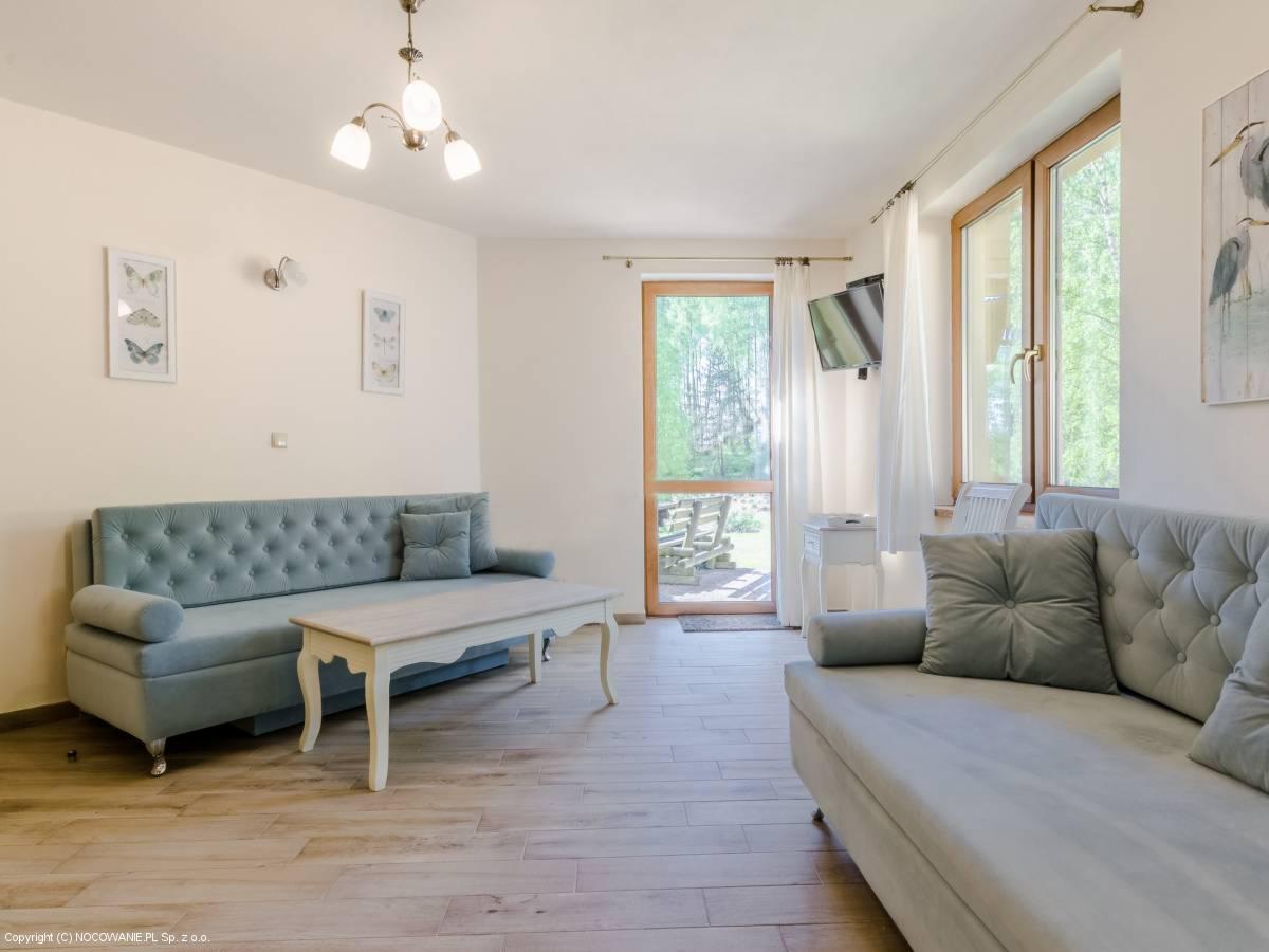 Jasne wnętrze salonu z dwoma turkusowymi kanapami, drewnianą podłogą, stolikiem kawowym i wyjściem na zewnątrz.