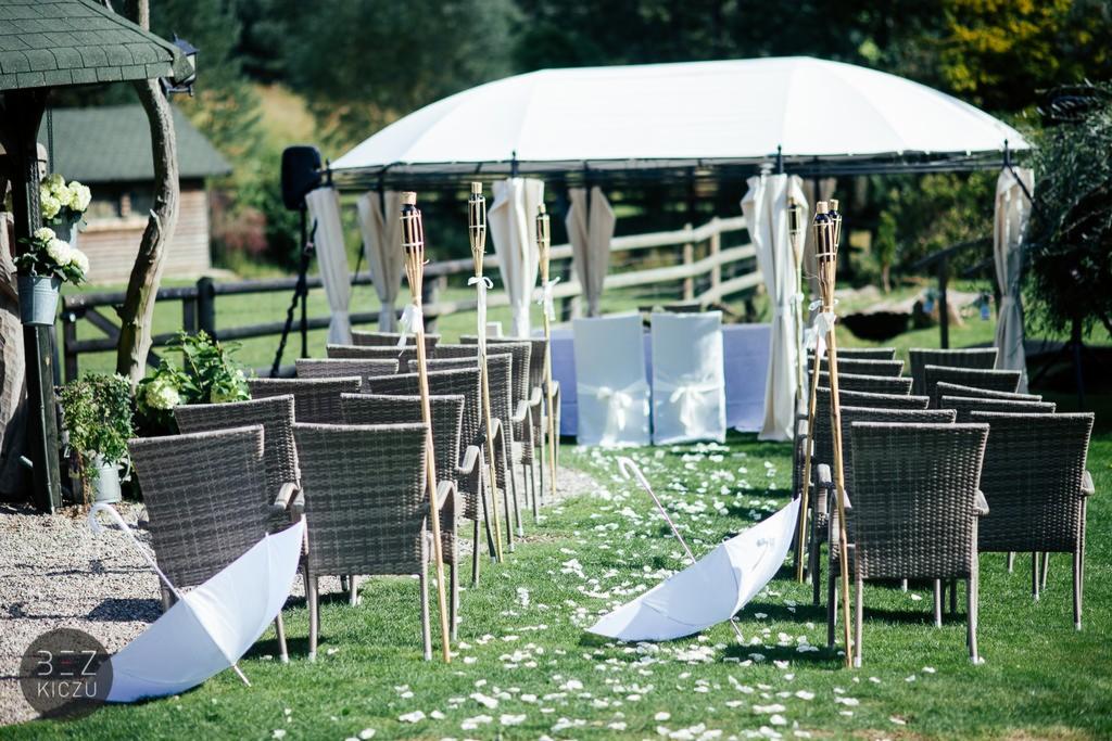 Przygotowania do ceremonii ślubnej na świeżym powietrzu: rzędy wiklinowych krzeseł, pochodnie tiki, biały baldachim, parasole i płatki kwiatów na trawniku.