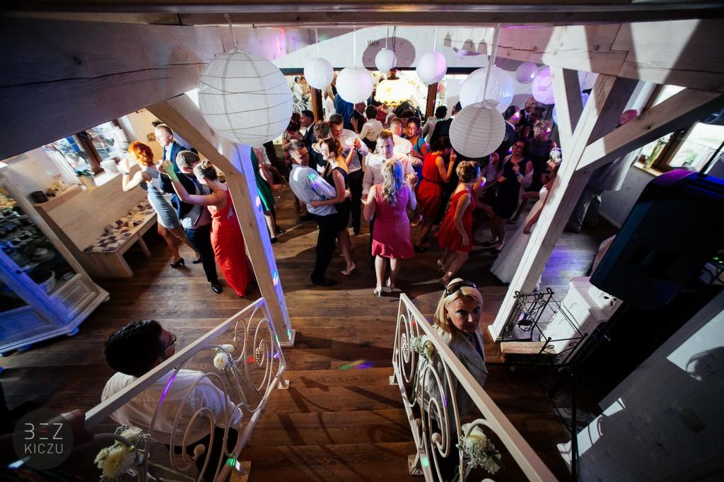 Sala weselna z drewnianym parkietem, białe lampiony, goście tańczący w tle, widok z góry ze schodów z ozdobną balustradą.