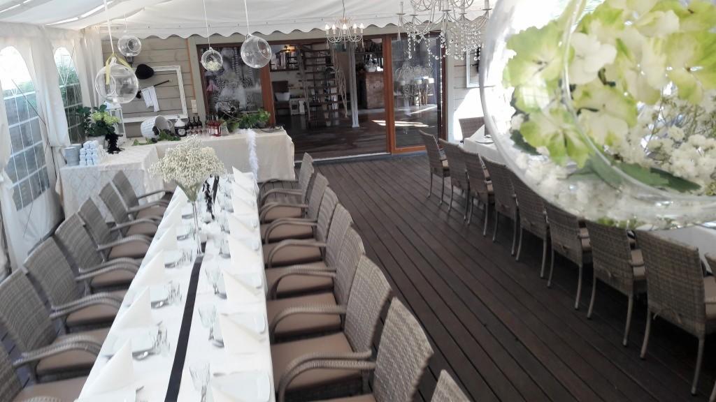 Uroczysta aranżacja sali w jasnych barwach z długim stołem, krzesłami z wikliny, kwiatowymi dekoracjami w szklanych kulach i kryształowymi żyrandolami pod białym namiotem.
