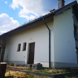 Nowo otynkowany dom jednorodzinny z ciemną stolarką okienną i drzwiową, widoczny fragment ogrodu z taczką i kostką brukową.