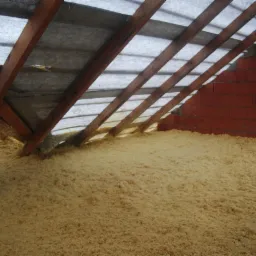 Poddasze w trakcie docieplania granulatem wełny, widoczne krokwie, membrana i fragment ściany z czerwonej cegły.