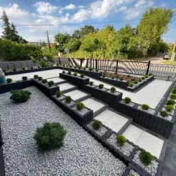 Nowoczesna aranżacja przestrzeni zewnętrznej z szarym żwirem, podwyższonymi rabatami z czarnych obrzeży, jasnymi płytami chodnikowymi i niskimi krzewami, widok na ogrodzenie i zieleń w tle.