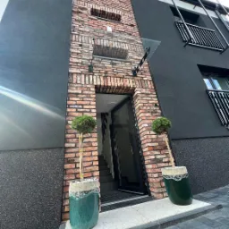 Nowoczesny budynek z elewacją w kolorze grafitowym i ceglanym, szklany daszek nad wejściem, dekoracyjne drzewka w ceramicznych donicach po bokach drzwi wejściowych.