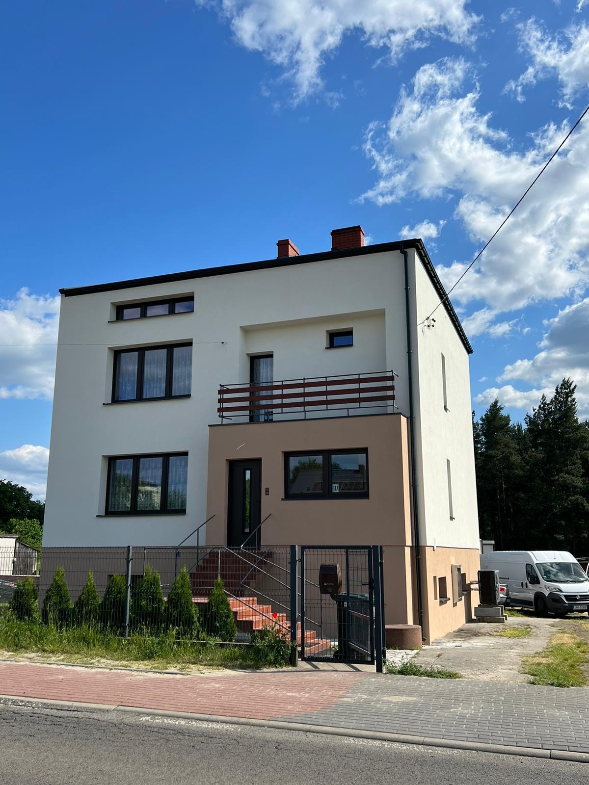 Dwukondygnacyjny dom z płaskim dachem, wykończony w bieli i beżu, z balkonem i schodami wejściowymi, ogrodzony metalową siatką.