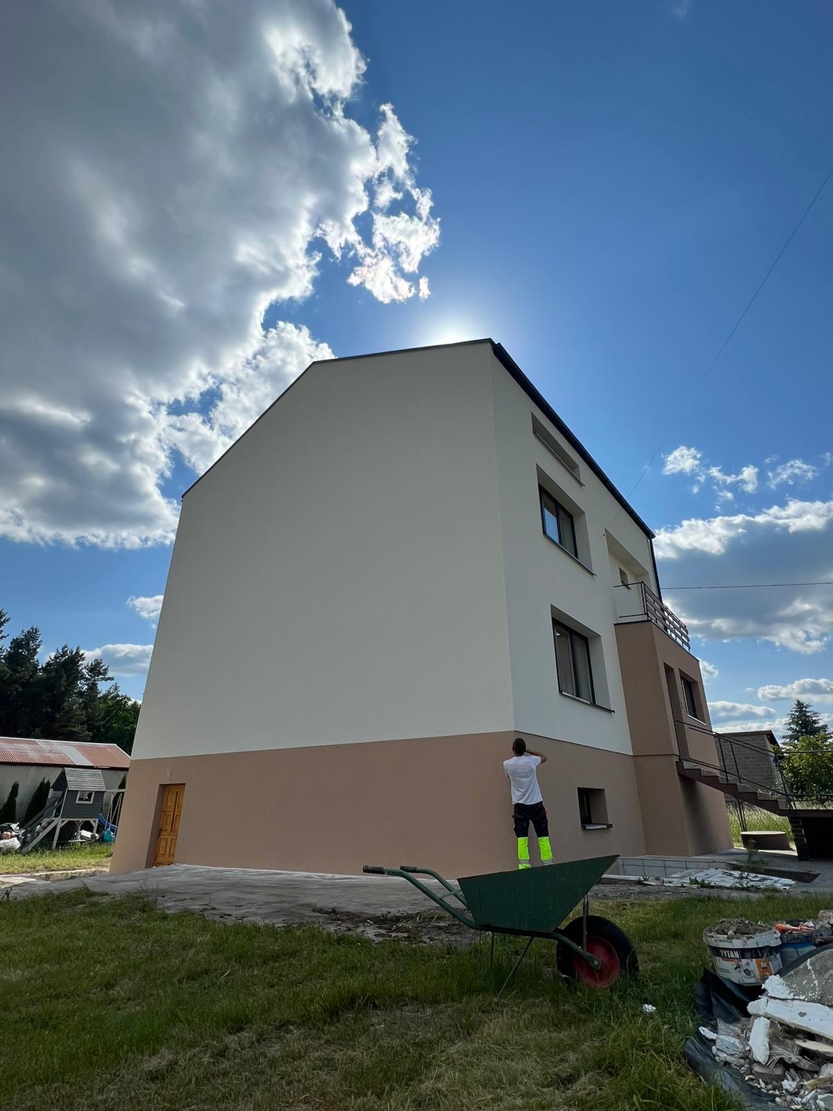 Dwukondygnacyjny budynek z elewacją w dwóch odcieniach beżu i bieli, pracownik w jaskrawych getrach wykańcza górną część elewacji, w tle błękitne niebo z chmurami i taczka na trawniku.