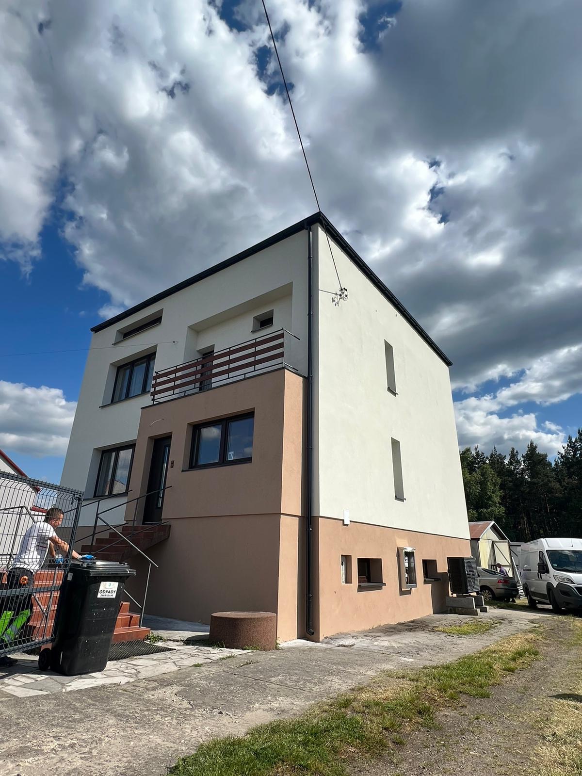 Dwukondygnacyjny dom po remoncie elewacji, z dwukolorową fasadą (beżowa dolna część i biała górna), balkonem z drewnianą balustradą oraz pracownikiem porządkującym teren przed budynkiem...