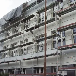 Budynek wielorodzinny w trakcie prac elewacyjnych, widoczne rusztowania i fragment zakrytej plandeką części ściany.