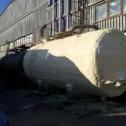Izolowany pianą zbiornik przemysłowy na zewnątrz budynku, widoczne ślady piany na ziemi i konstrukcji wsporczej.