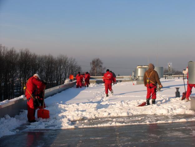 Grupa pracowników w czerwonych kombinezonach odśnieża płaski dach budynku, używając łopat. W tle widoczne drzewa i elementy wentylacji.