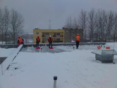 Pracownicy w pomarańczowych kamizelkach odblaskowych odśnieżają płaski dach budynku, używając czerwonych łopat. W tle widoczny żółty blok mieszkalny i nagie drzewa.