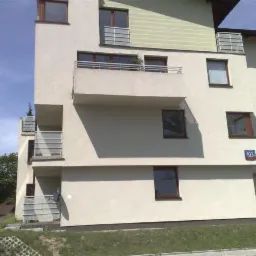 Nowoczesny budynek wielorodzinny z jasną elewacją, balkonami z metalowymi barierkami i numerem 103 na ścianie, widoczny fragment zielonego dachu i trawnik.