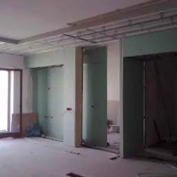 Wnętrze w trakcie remontu: montaż stelaża sufitowego i płyt kartonowo-gipsowych na ścianach działowych, widoczne instalacje elektryczne i rury na podłodze, okno balkonowe z ciemnymi ramami.