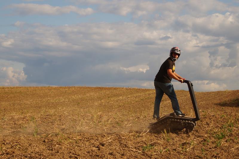 Mężczyzna w kasku jeżdżący na gąsienicowym pojeździe DTV Shredder po zaoranym polu, widok z boku, pochmurne niebo w tle.