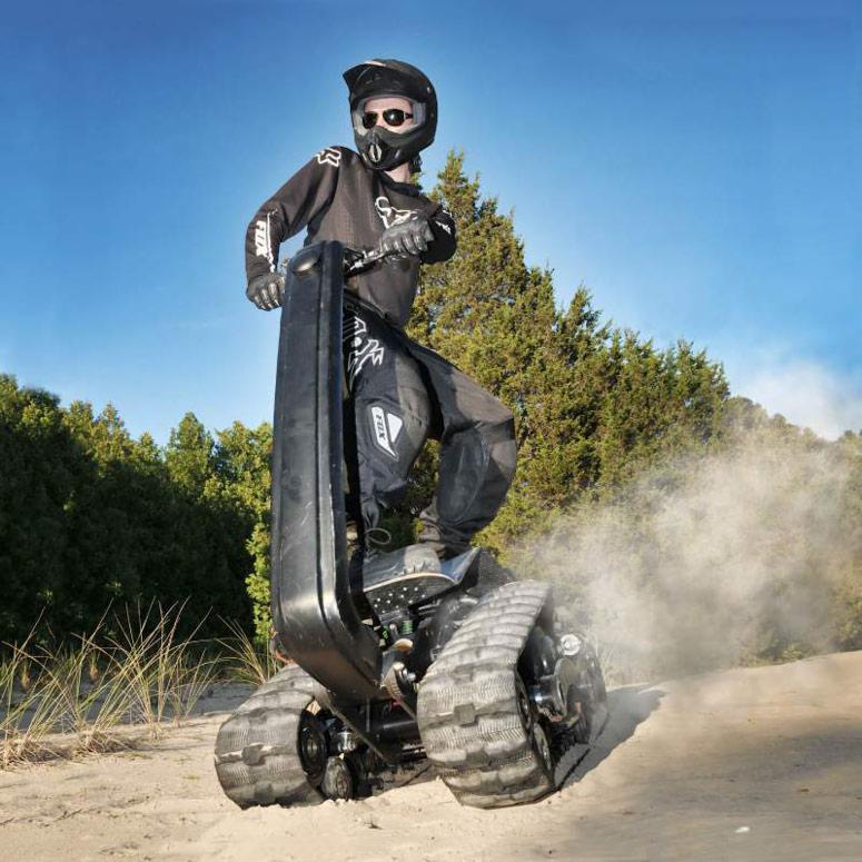 Osoba w kasku i kombinezonie na pojeździe gąsienicowym w terenie piaszczystym, z drzewami w tle.