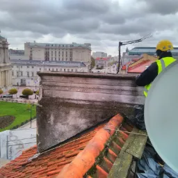 Pracownik w kasku i kamizelce odblaskowej na dachu z czerwonej dachówki, antena satelitarna w tle, widok na miasto w pochmurny dzień.