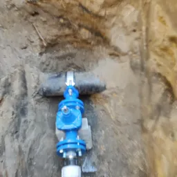 Niebieski zawór na rurze w wykopie ziemnym, zabezpieczony metalową obejmą.