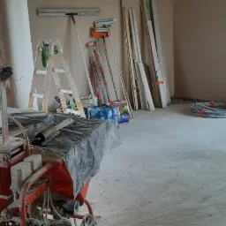 Wnętrze w trakcie prac wykończeniowych, z maszyną do nakładania tynków na pierwszym planie, drabiny opierające się o ścianę, oraz zwój kabli przy oknie.