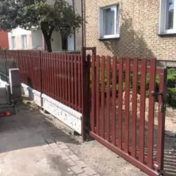 Ciemnobrązowe ogrodzenie z pionowych paneli z furtką, zamontowane na niskim, betonowym murku, na tle bloku mieszkalnego z cegły.