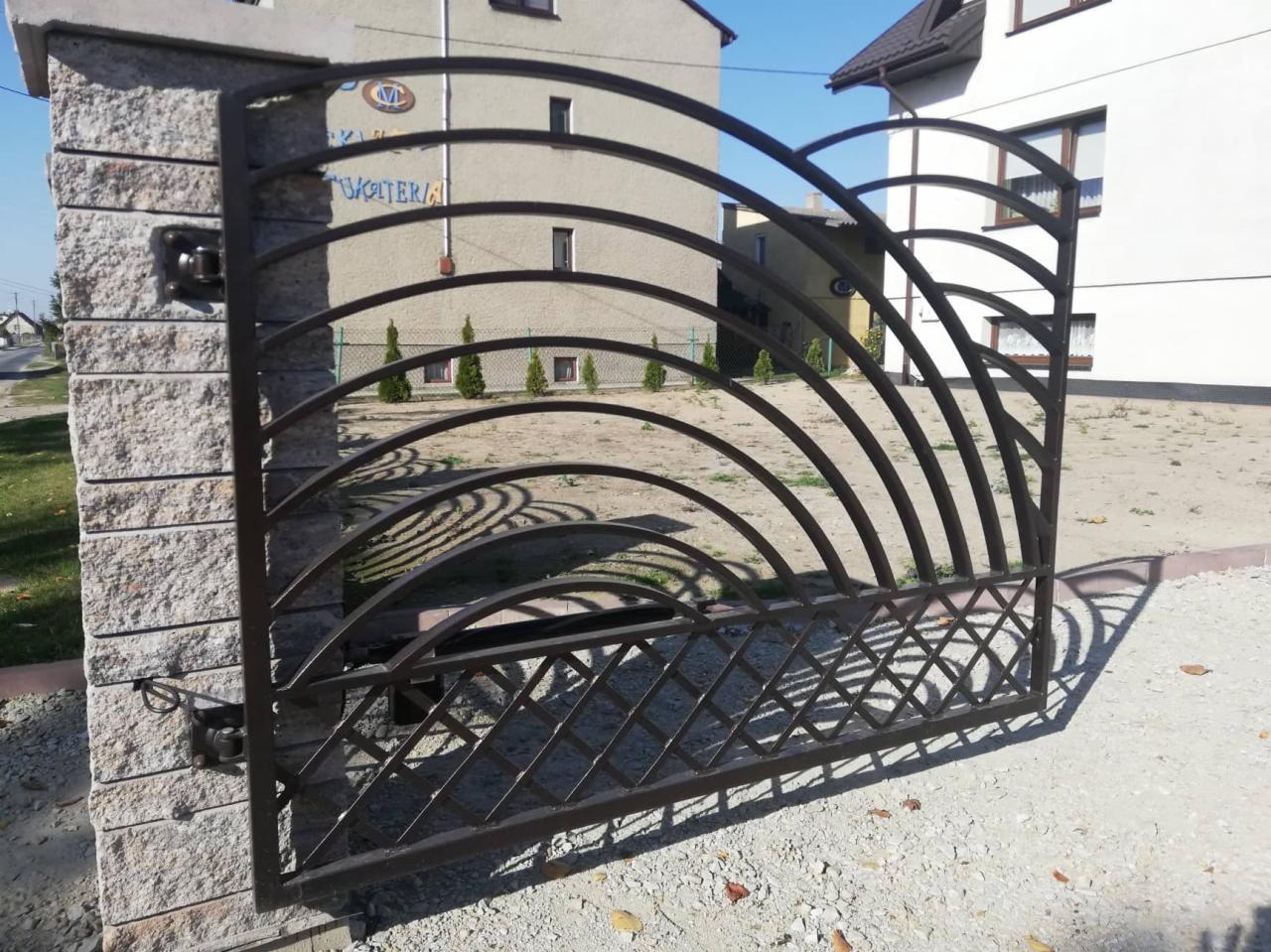 Metalowa brama wjazdowa z łukowatymi elementami, zamontowana na kamiennych słupkach, na tle budynków mieszkalnych i zieleni.