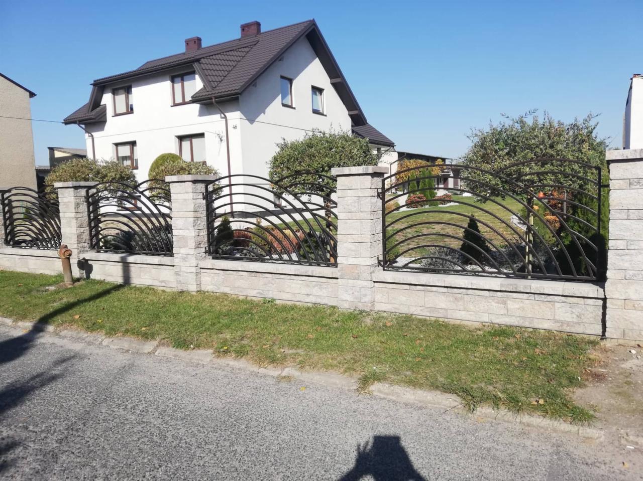 Czarny metalowy płot z łukowatymi przęsłami i kamiennymi słupkami, na tle dwupiętrowego domu z brązowym dachem i zielenią.