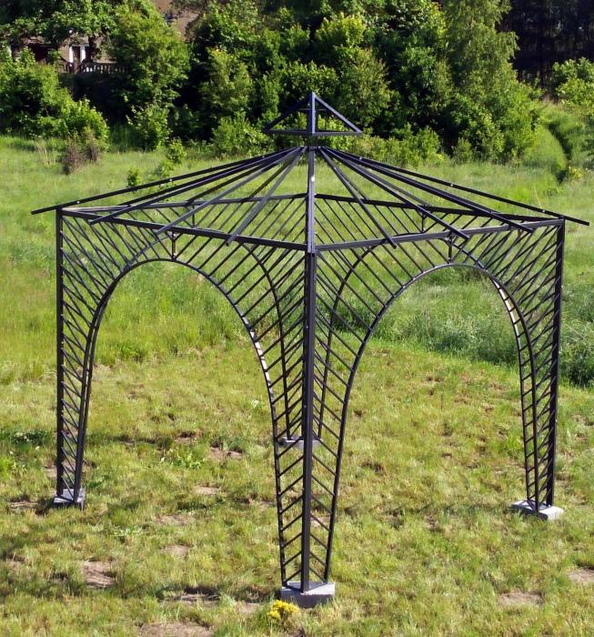 Czarna, metalowa altana ogrodowa o konstrukcji łukowej, z ukośnymi prętami, ustawiona na trawniku z widokiem na drzewa i budynek w tle.