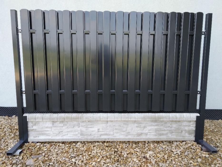 Fragment nowoczesnego ogrodzenia panelowego w kolorze grafitowym, osadzonego na betonowej podmurówce imitującej kamień, widoczny na tle jasnej ściany i żwirowego podłoża.