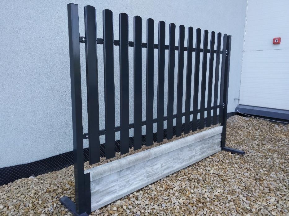 Fragment czarnego ogrodzenia panelowego z podmurówką imitującą kamień, ustawiony na żwirowym podłożu przed budynkiem z szarą elewacją i białą bramą garażową.
