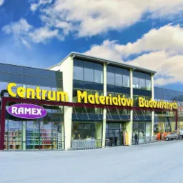 Budynek Centrum Materiałów Budowlanych RamEX z dużymi oknami i żółtym napisem na tle błękitnego nieba z białymi chmurami.