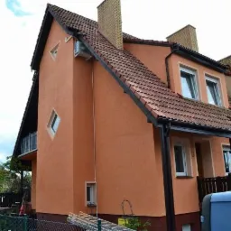 Pomarańczowa elewacja szeregowca z klimatyzatorem i czarnymi rynnami, widok z dołu, drabina oparta o ścianę. Dachówka ceramiczna.