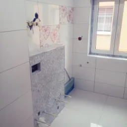 Biała łazienka w trakcie wykańczania: świeżo położone płytki z kwiatowym motywem, obudowa stelaża WC z mozaiki, okno z widokiem na budynek.