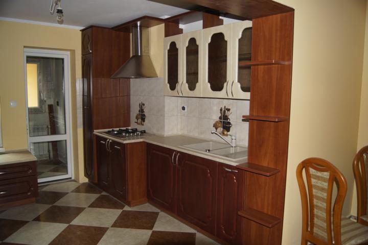 Kuchenna zabudowa w odcieniach brązu i beżu, z płytek ceramicznych na ścianie z motywem przyborów kuchennych, widoczny fragment blatu i zlewu oraz fragment krzesła na pierwszym planie.