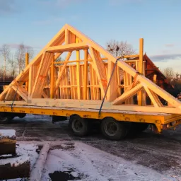 Gotowe elementy konstrukcji drewnianej dachu, zabezpieczone pasami transportowymi, przewożone na żółtej przyczepie w zimowej scenerii.