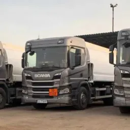 Trzy szare cysterny Scania z pomarańczowymi tablicami ostrzegawczymi, zaparkowane obok siebie na placu.