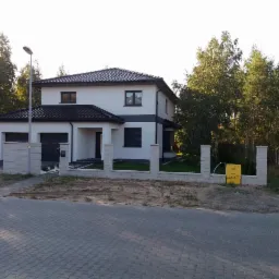 Dwukondygnacyjny dom z garażem i ciemną dachówką, ogrodzony niskim, murowanym płotem z elementami dekoracyjnymi, na tle drzew.