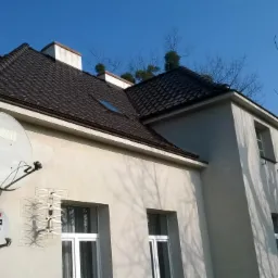 Elewacja dwukondygnacyjnego budynku z ciemnobrązowym dachem, widoczne dwie anteny satelitarne i antena telewizyjna na ścianie, drzewa bez liści w tle, błękitne niebo.