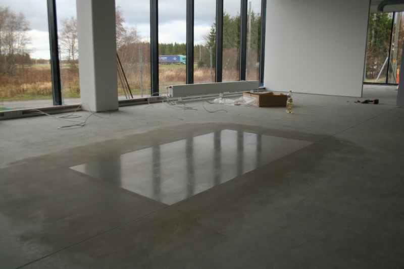 Fragment betonowej podłogi po nabłyszczaniu, z wyraźnym odbiciem okien, widoczne kable i kartonowe pudło w tle.