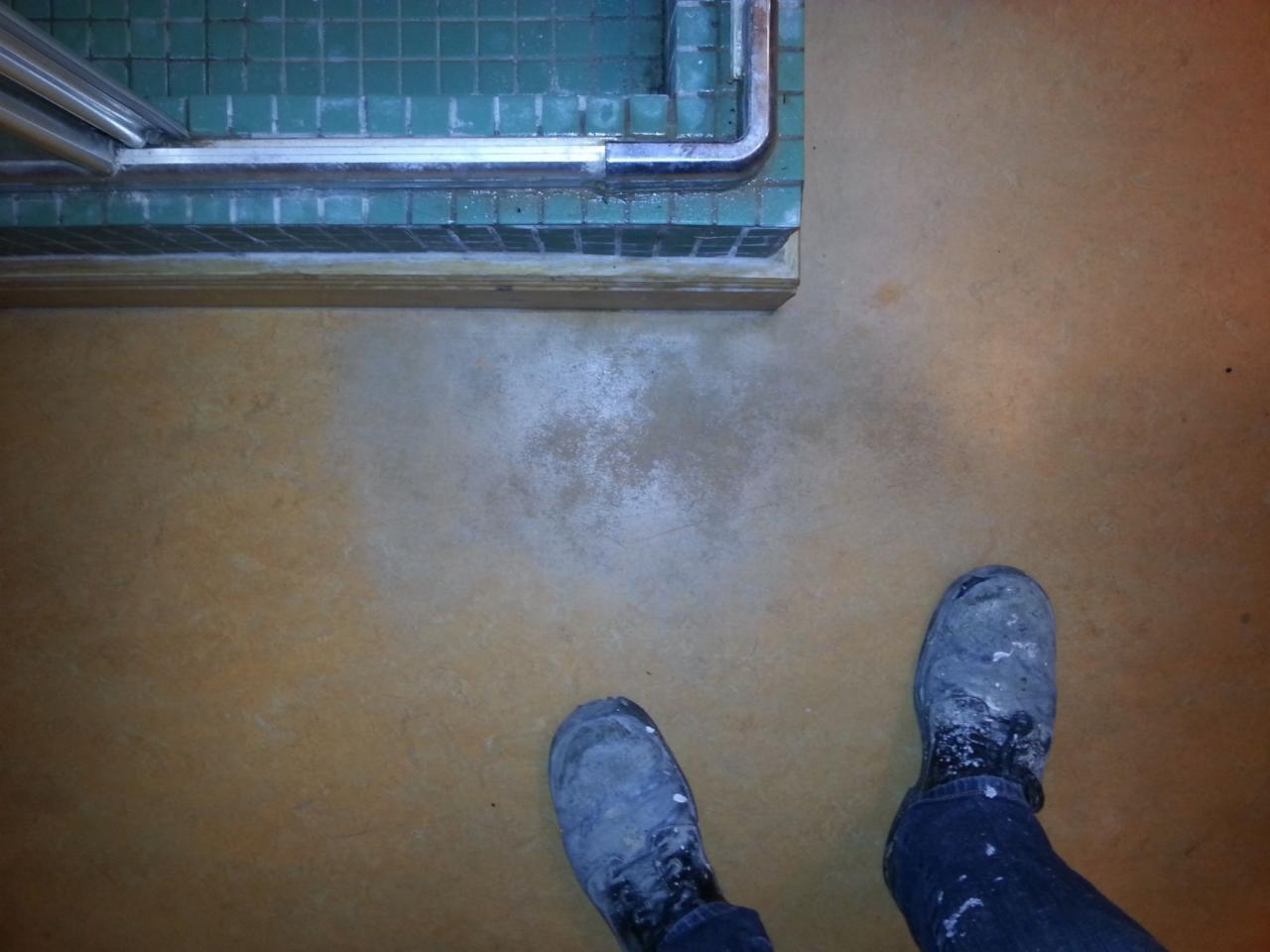 Podłoga z PCV/linoleum w trakcie renowacji, widoczne ślady po szlifowaniu i buty robocze z zabrudzeniami farby, fragment kabiny prysznicowej z zielonymi płytkami w tle.