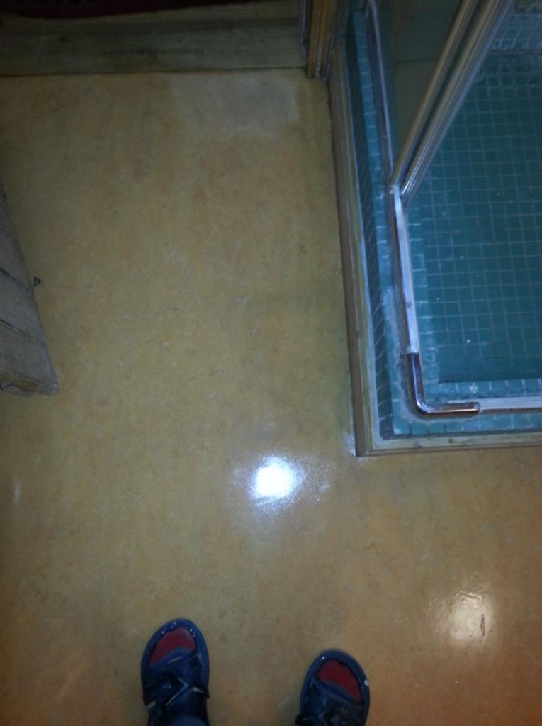 Odnowiona podłoga z PCV o jednolitej, jasnobrązowej barwie, widoczne odbicie światła, fragment kabiny prysznicowej z boku, ujęcie z góry na stopy w sandałach.