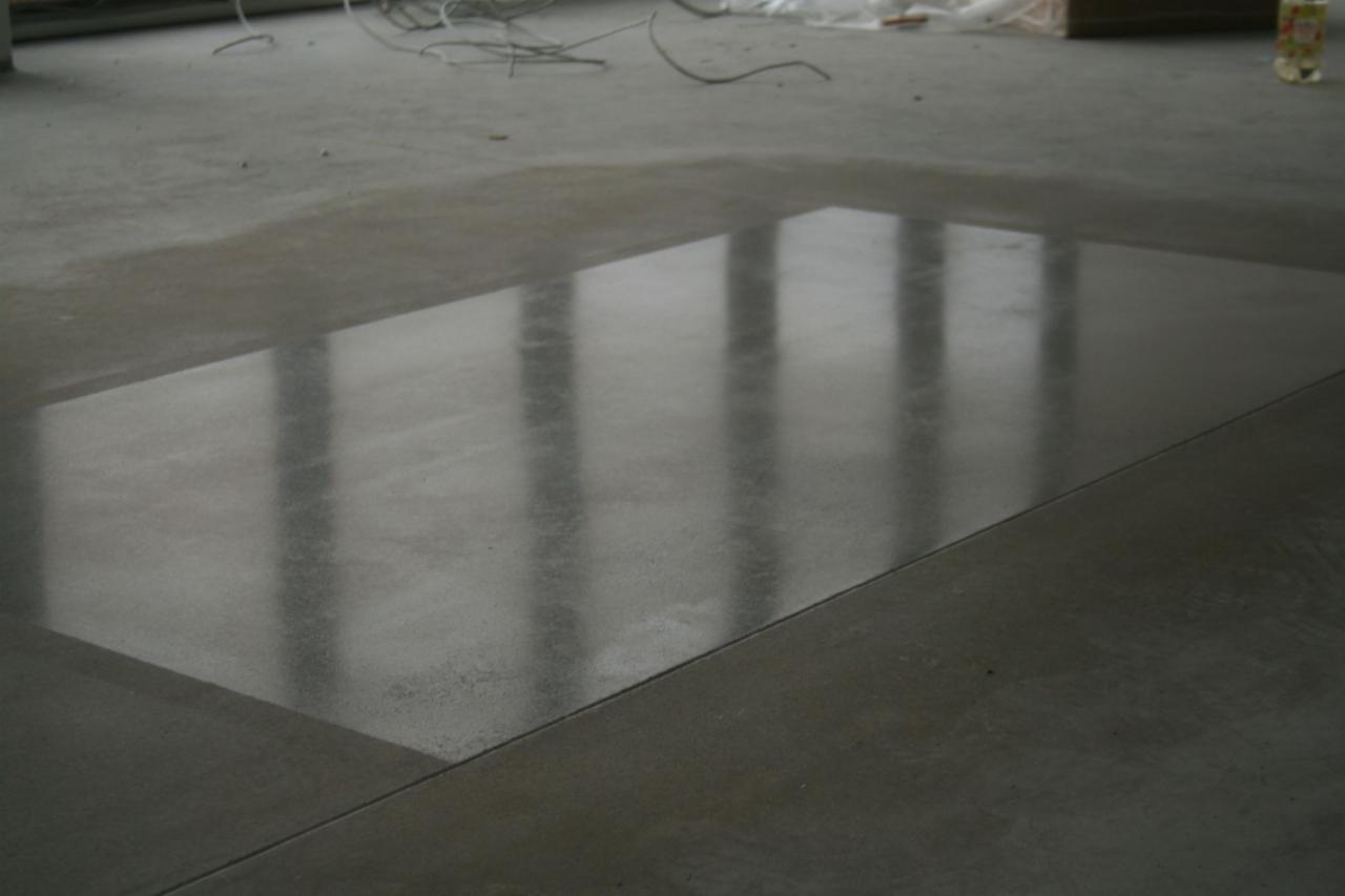 Wypolerowana, betonowa podłoga z wyraźnym odbiciem okien, widoczne kable i materiały budowlane w tle.