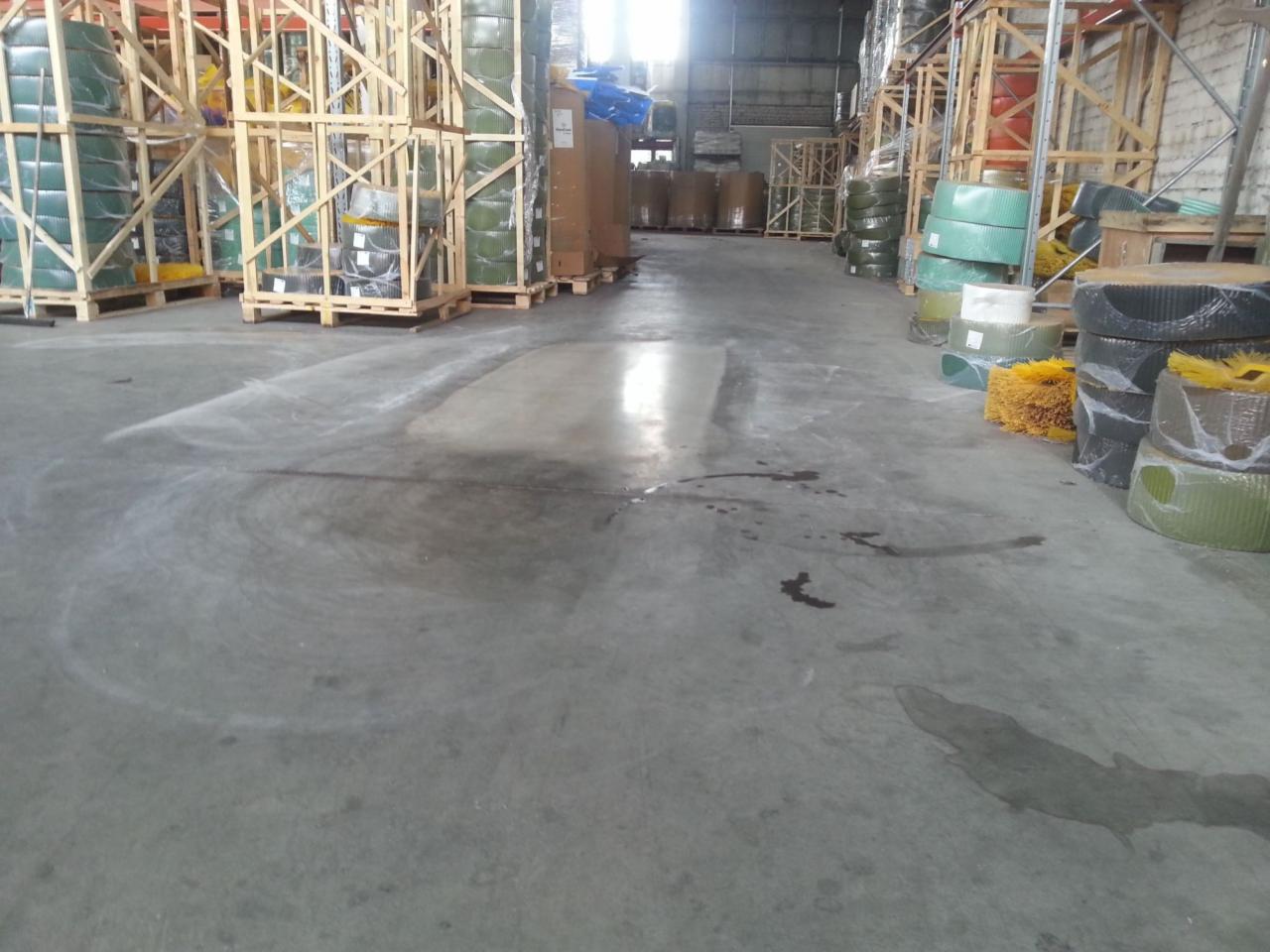 Szlifowana posadzka betonowa w hali magazynowej, widoczne ślady po procesie renowacji i plamy, w tle regały z towarem.