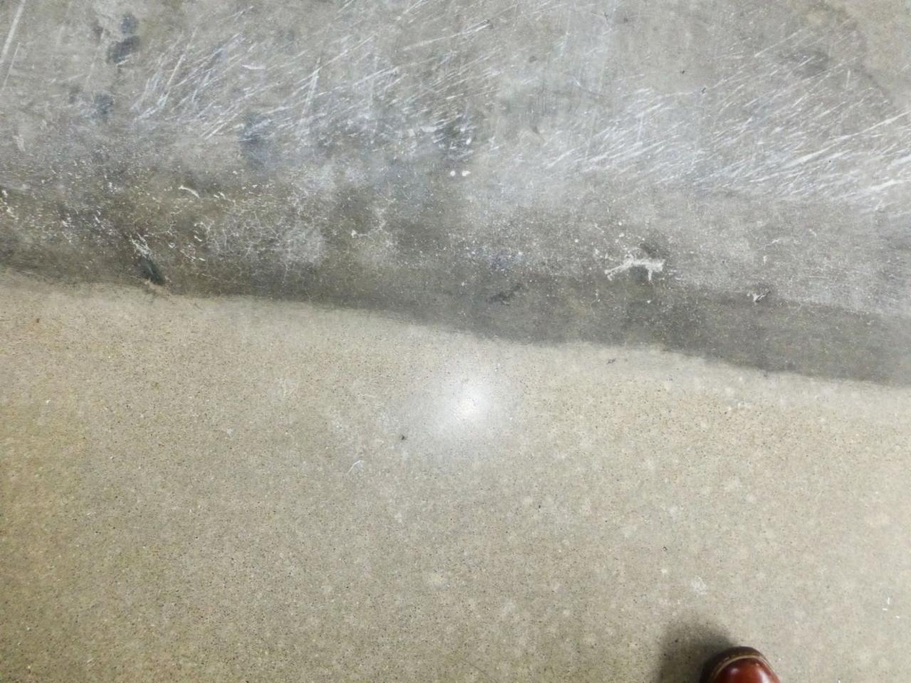 Fragment betonowej podłogi w trakcie renowacji, widoczna różnica w kolorze i fakturze pomiędzy odnowioną i nieodnowioną częścią, z fragmentem brązowego buta w rogu kadru.