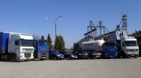 Szeroki kadr na flotę pojazdów (ciężarówki i samochody osobowe) przed składem budowlanym z silosami i zbiornikiem, w słoneczny dzień.