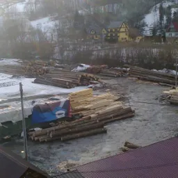 Widok z góry na plac tartaku w zimowej scenerii: stosy okrąglaków i tarcicy, częściowo przykryte śniegiem, w tle zabudowania i zalesione wzgórza.